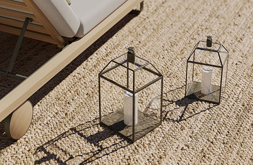 Close-up of a wooden sun lounger with cream cushions beside a pool, with two decorative metal lanterns on a gravel terrace.