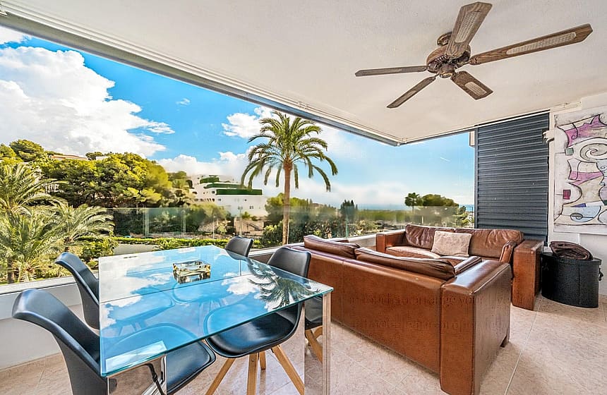 A covered terrace with a glass dining table, leather sofa, and ceiling fan overlooking a coastal landscape with palm trees.