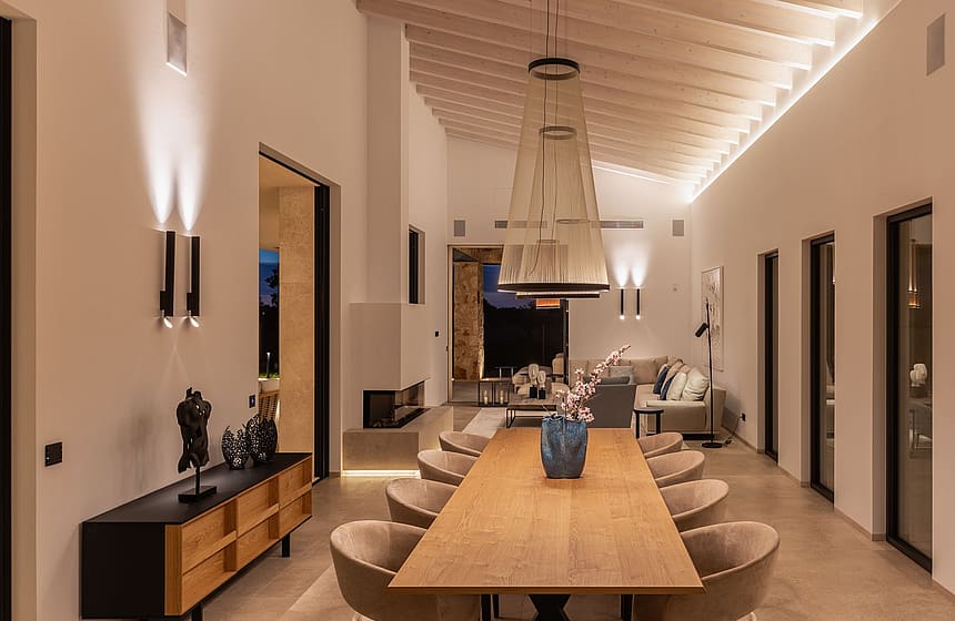Dining room with a long oak table and curved chairs, featuring a high gabled ceiling with exposed beams and recessed lighting.