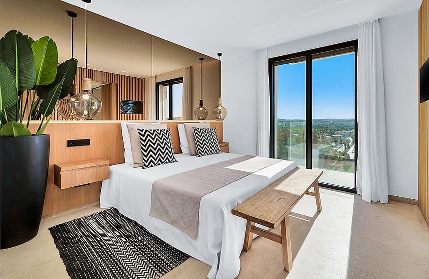 A modern bedroom featuring a bed with a wooden headboard, a large mirror, a wooden bench, and a floor-to-ceiling glass door.
