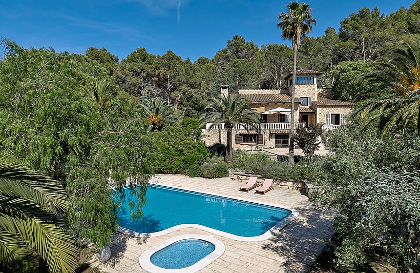 Exterior view of a traditional stone finca surrounded by trees, featuring a rectangular swimming pool and a separate plunge pool on a stone patio.