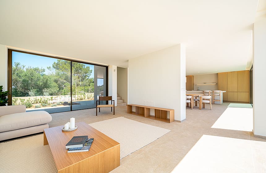 A bright, open-plan living room with a light beige sofa, wooden coffee table, and large sliding glass doors overlooking trees.