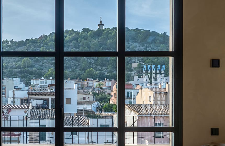A bedroom featuring a large grid-style window overlooking a Mediterranean town and green hillside.