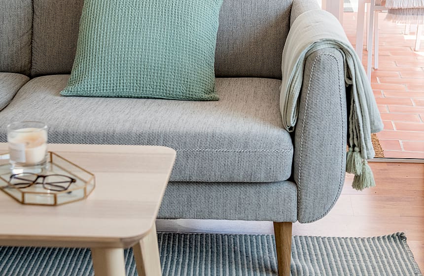 Close-up of a grey woven sofa with a mint green cushion and a light wood coffee table on a striped rug, leading to a terrace.