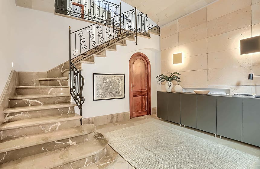 An interior shot of an entrance hall featuring a wide marble staircase with a wrought-iron railing, a curved wooden door, and a textured stone feature wall.