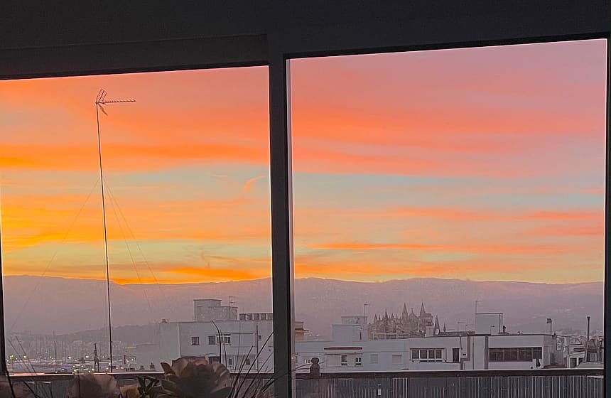View through a glass door showing an urban skyline at sunset, with a silhouette of an outdoor dining area on the terrace.