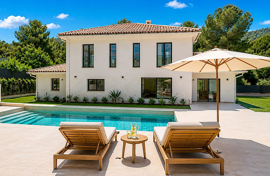 Exterior view of a two-story white villa with clay-tiled roof, rectangular pool, and sun loungers on the paved terrace.