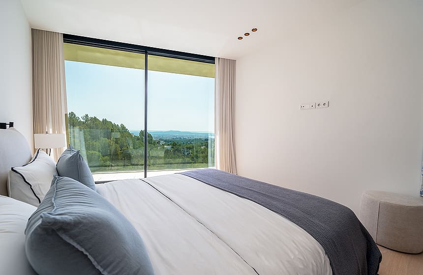 Modern bedroom with a large bed, floor-to-ceiling sliding glass doors, and a distant sea view framed by pine trees.