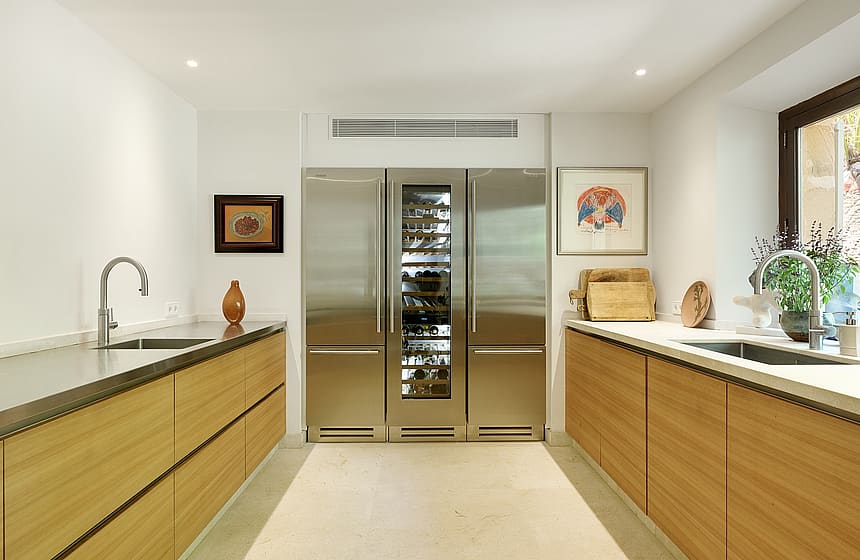 A modern galley kitchen featuring oak cabinetry, stainless steel appliances, and a central glass-fronted wine fridge.