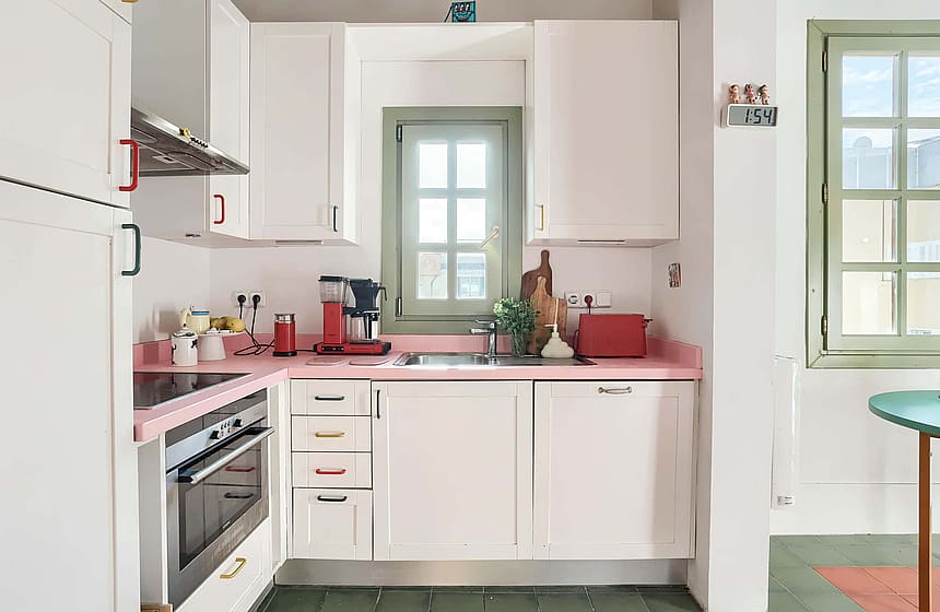 A compact kitchen with white cabinetry, pink stone surfaces, a stainless steel oven, and olive green window frames.