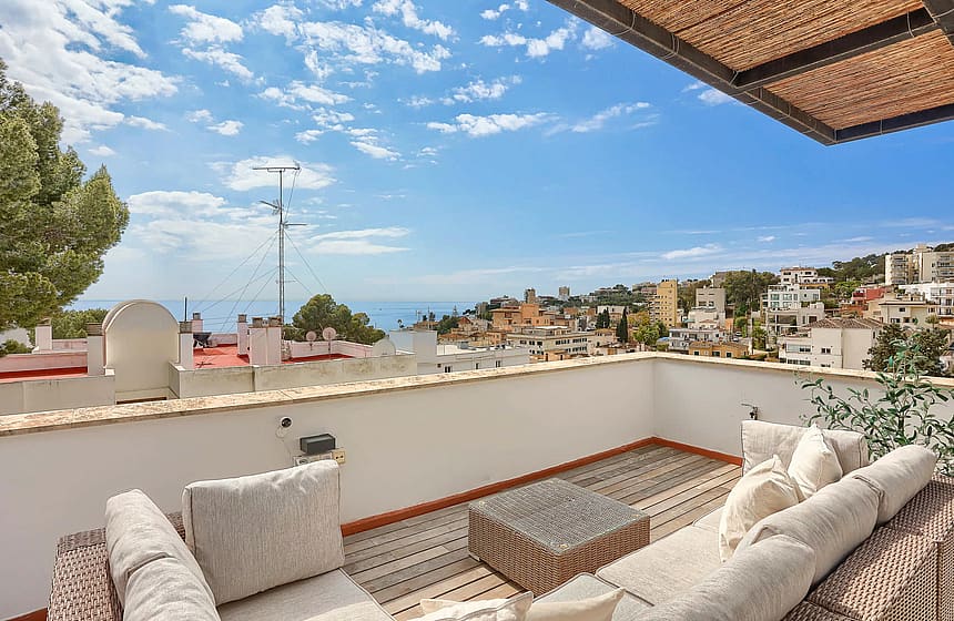 A rooftop terrace featuring a modular sofa and coffee table under a bamboo shade, overlooking coastal houses and the ocean.