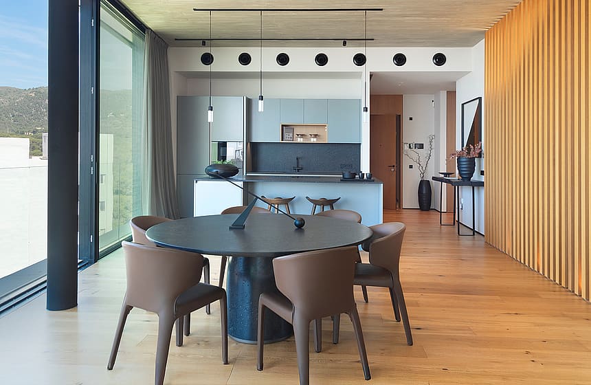 Modern open-plan kitchen with a dark circular dining table, leather chairs, and wooden slat wall paneling, featuring floor-to-ceiling sliding glass doors.