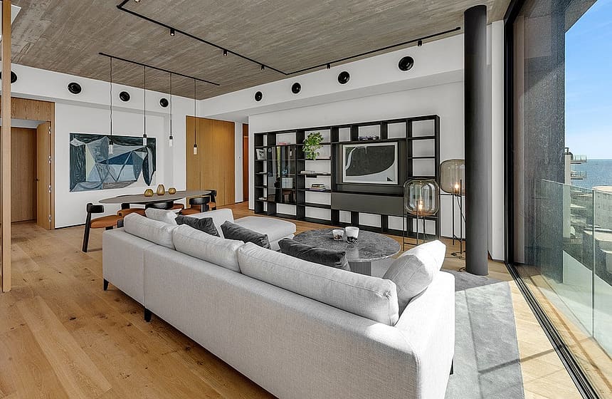 Modern open-plan living room with light oak flooring, concrete ceiling, track lighting, a large light gray sofa, and full-height glass walls overlooking the sea.