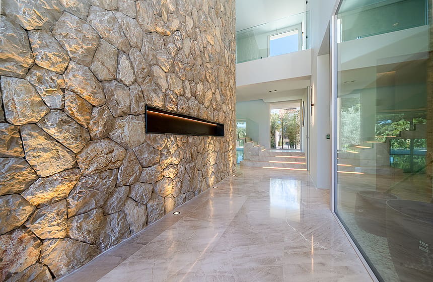 Interior entrance hall with a large dry-stone feature wall, linear fireplace, polished light marble floors, and glass partitions.