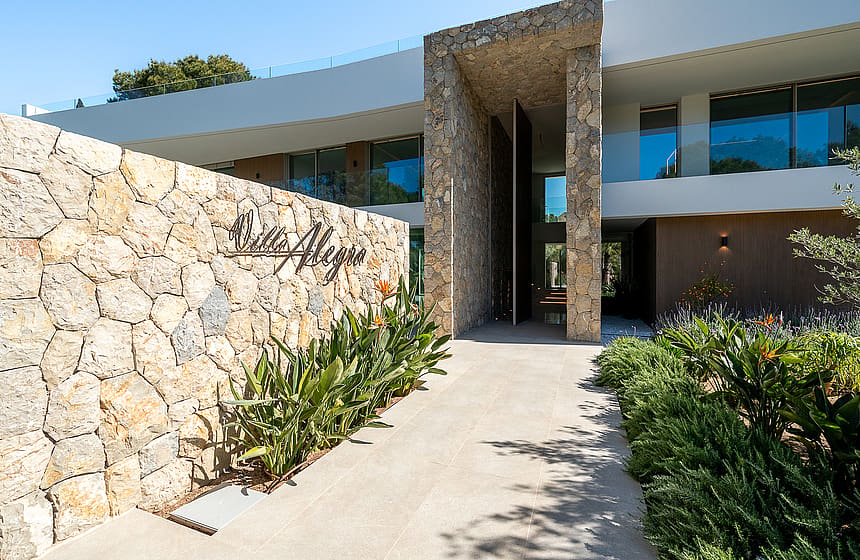 Modern villa entrance featuring a curved natural stone retaining wall, paved walkway, and landscaping in Santa Ponsa.