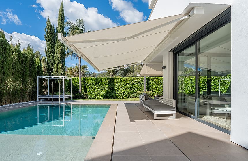 Heated saltwater swimming pool with sun loungers, retractable awning and tall hedge screening.