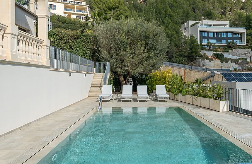 An outdoor rectangular swimming pool with four white loungers on a stone-paved terrace, bordered by a white villa wall.