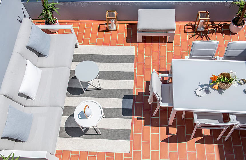 Aerial view of a tiled penthouse terrace featuring a gray sectional sofa, a white dining table with chairs, and a striped rug.