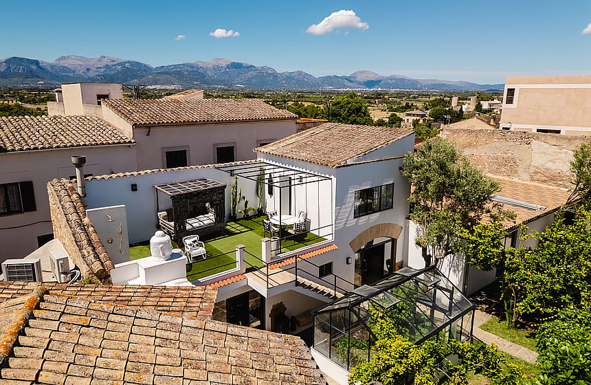 High-angle view of a townhouse exterior with a sunlit rooftop terrace featuring artificial grass, outdoor seating, and a pergola, set against a backdrop of distant mountains.