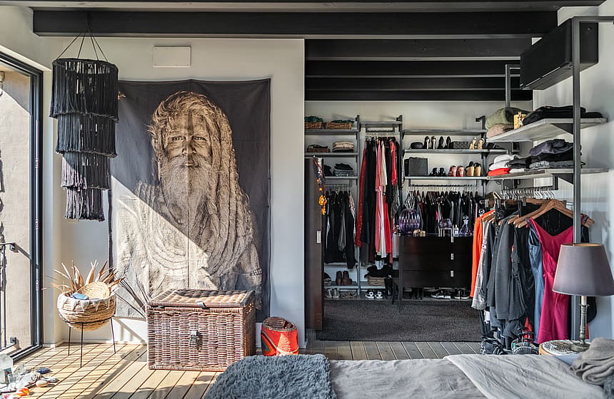 Walk-in wardrobe with industrial open shelving, dark wooden trunk, and decorative wall tapestry, illuminated by natural light.