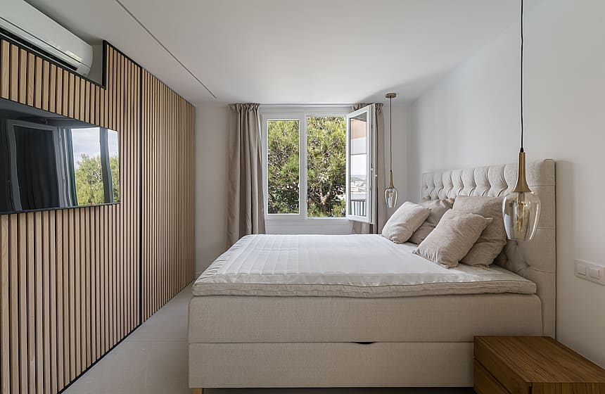 Master bedroom featuring vertical light wood wall panels, a wall-mounted television, and windows overlooking green trees.