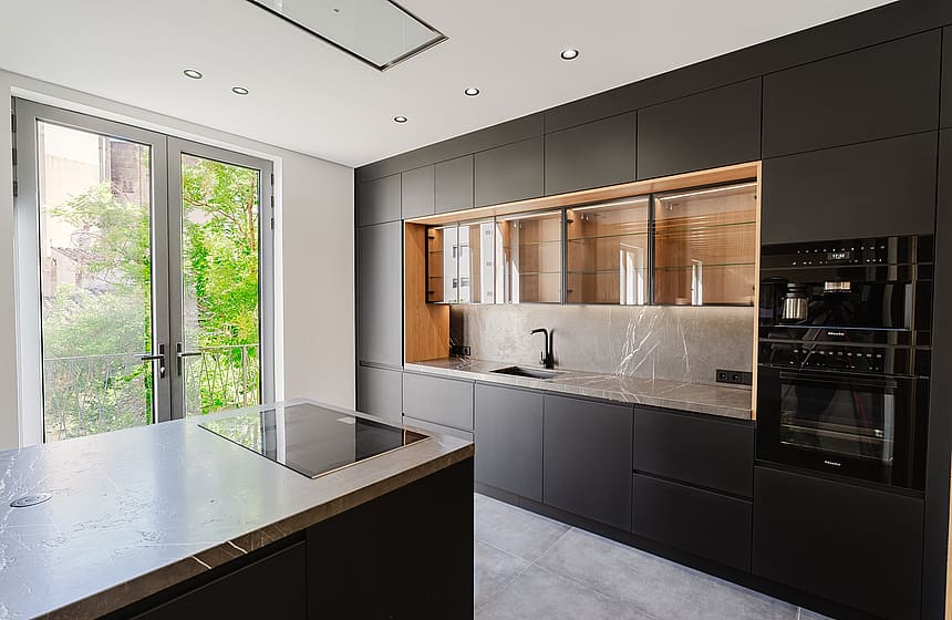 A modern kitchen with matte black cabinetry, a central island with an integrated hob, and French doors opening to greenery.