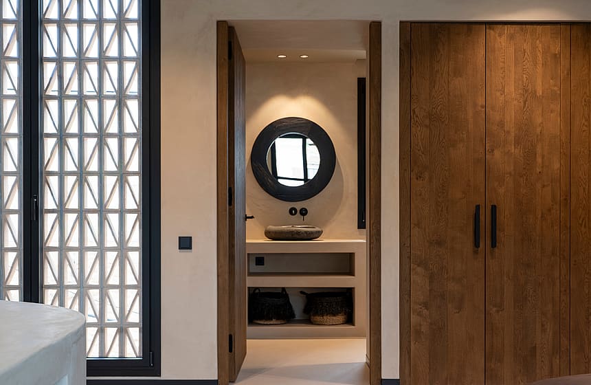 View from a bedroom into a modern bathroom with a circular mirror, stone basin, and wooden wardrobe doors on the right.