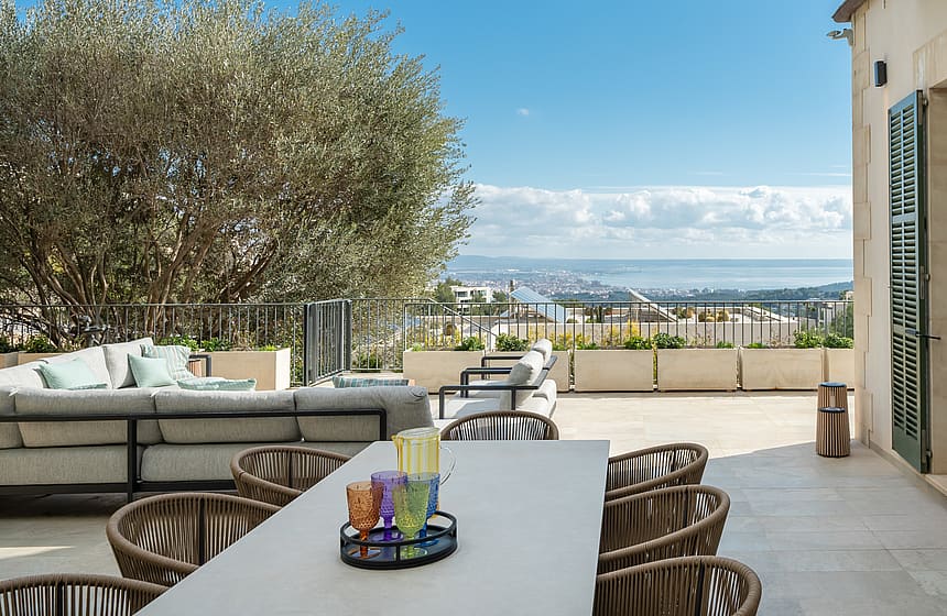 Outdoor dining terrace featuring a stone table, woven chairs, and a large olive tree overlooking the Son Vida landscape.