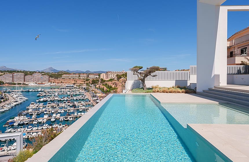 A modern infinity pool on a white terrace overlooking a busy marina and the sea under a clear blue sky.