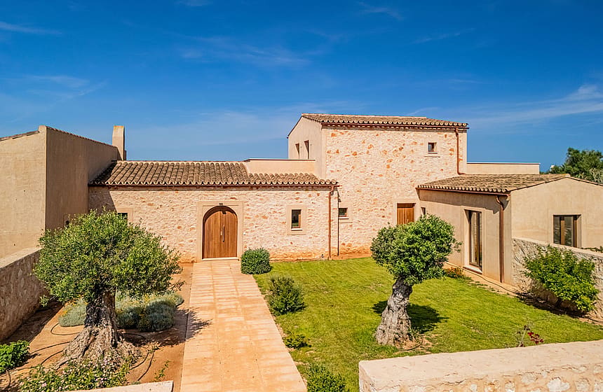 Exterior view of a traditional stone finca in Santanyí with a paved pathway leading to a wooden arched door, surrounded by a lawn and olive trees under a bright blue sky.
