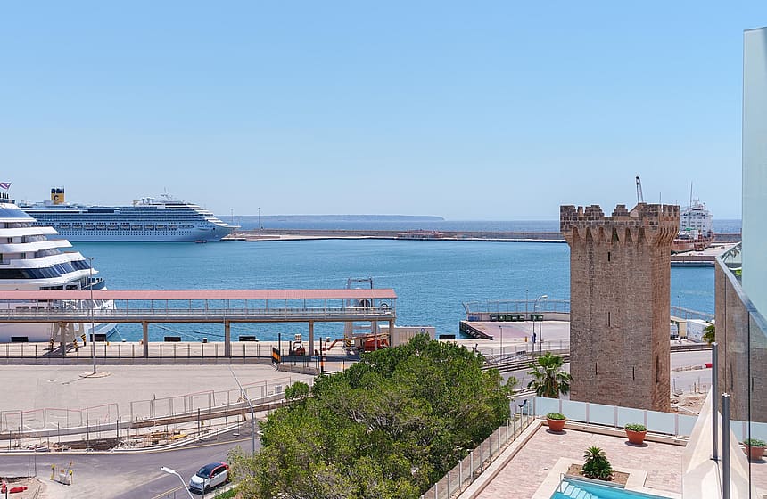 Vista elevada sobre un puerto con una torre histórica, cruceros y el mar bajo un cielo despejado.