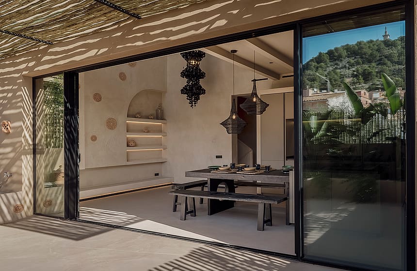 An open-plan dining room with a long wooden table and benches leading onto a terrace shaded by a reed canopy.