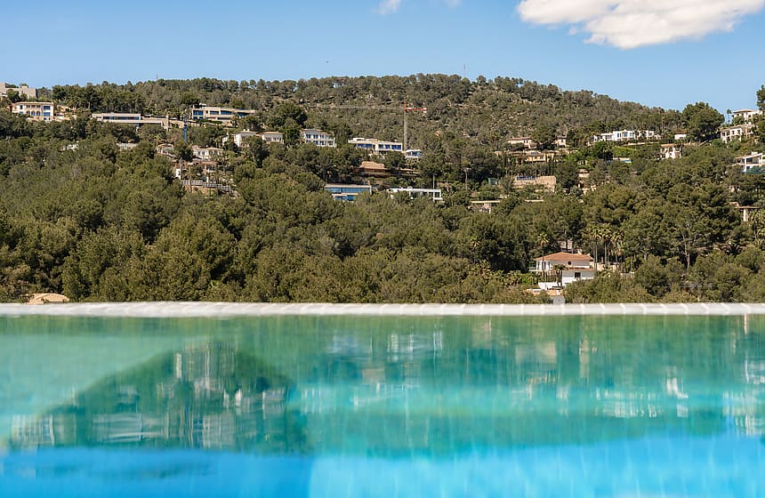 Close-up of an infinity pool edge reflecting the lush green hillside and villas of Son Vida under a clear sky.