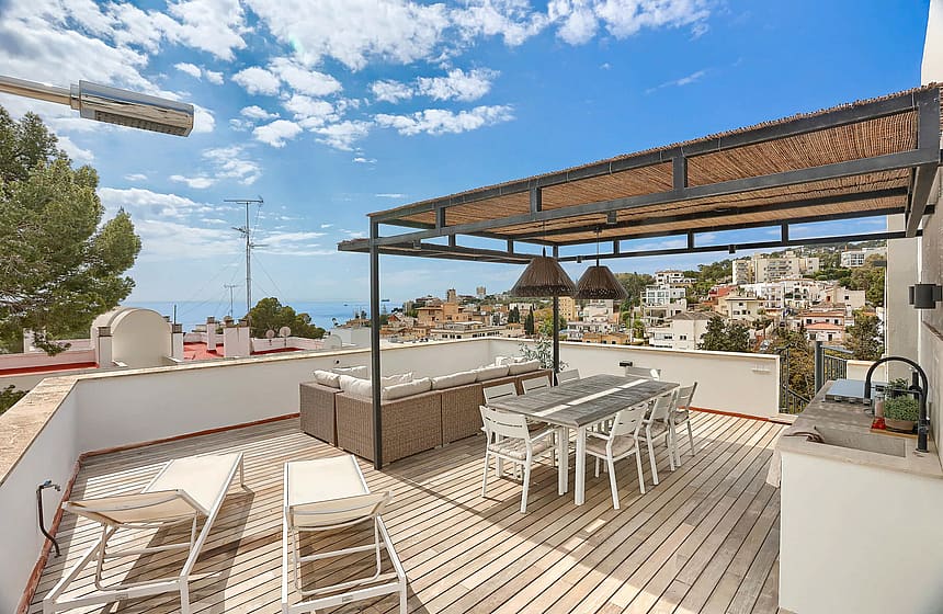A spacious outdoor rooftop terrace featuring a wooden deck, a covered dining area with a woven pergola, lounge seating, and distant sea views over a coastal neighborhood.