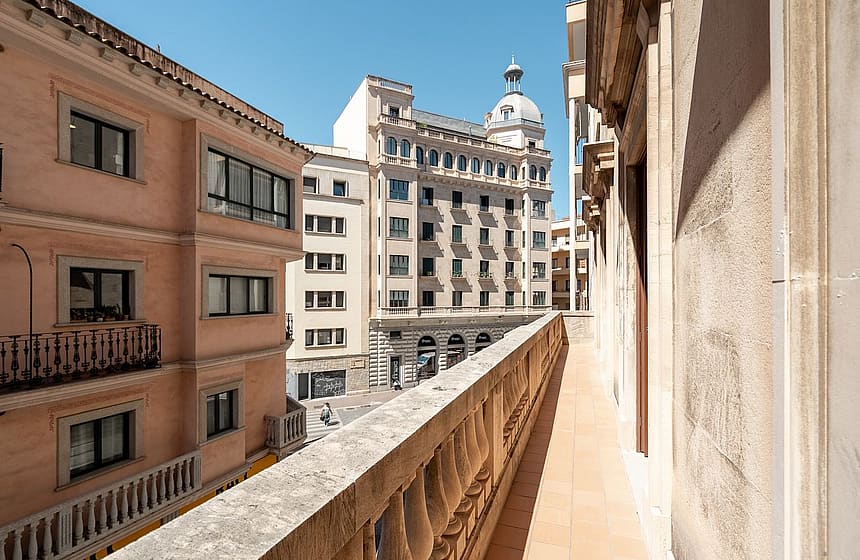 A stone terrace balcony with a classical balustrade overlooking a historic urban street with multi-story buildings in Palma.