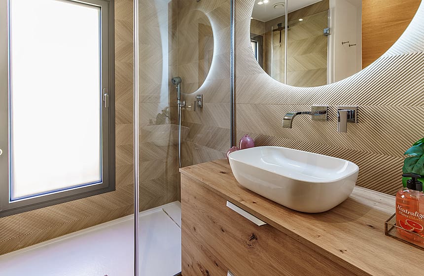 Modern bathroom featuring patterned beige wall tiles, a wooden vanity with a white vessel sink, and a glass-enclosed shower.