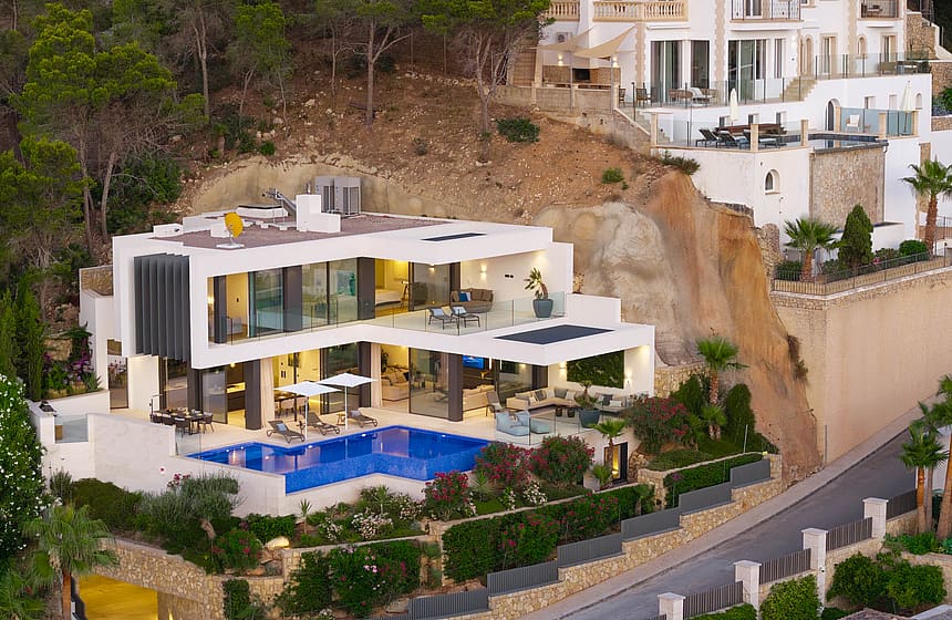 Aerial view of a white modern villa carved into a cliffside, featuring a swimming pool, multiple terraces, and glass facade.