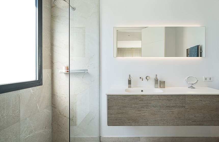 A modern bathroom featuring a glass-enclosed shower with light-toned stone tiling, a floating vanity with a wooden cabinet, and a wall-mounted mirror.