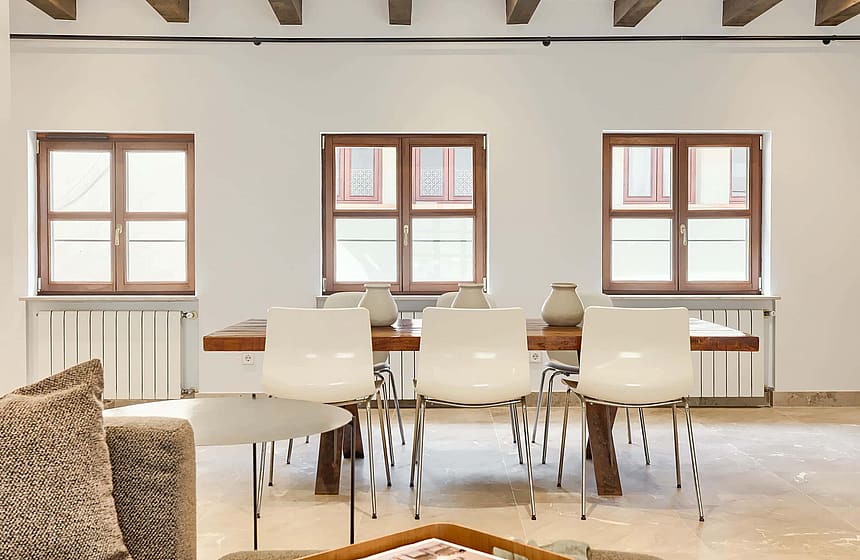Dining room with a wooden table, white modern chairs, and three wood-framed windows looking out to the city.
