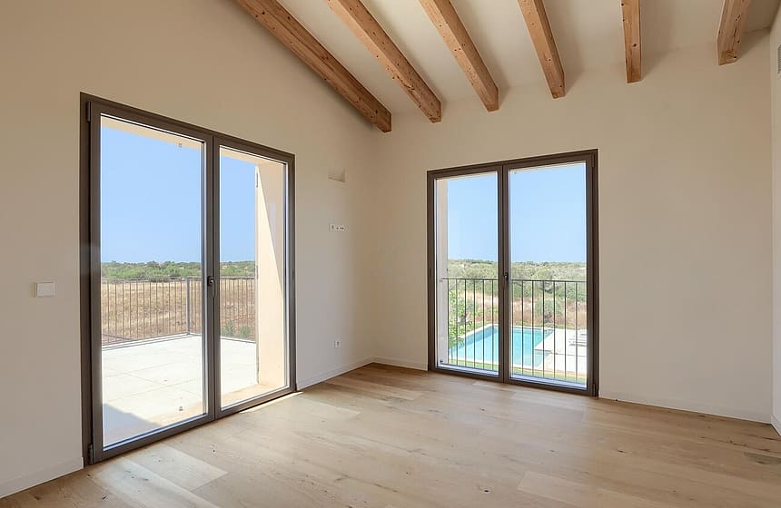 An unfurnished bedroom in a Finca, featuring light oak wood floors, white walls, exposed wooden ceiling beams, and sliding glass doors opening to a view of the pool and surrounding landscape.
