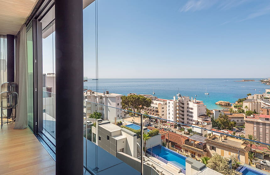 A glass-railed balcony looking out over the urban coastline of Cala Mayor with a swimming pool visible below.