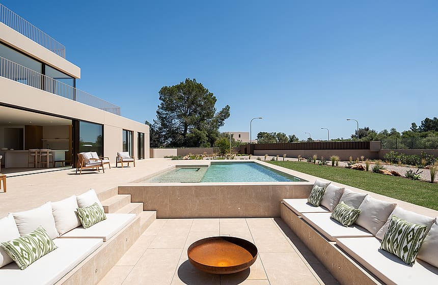 A sunken outdoor seating area with stone benches and white cushions faces a rectangular swimming pool under a blue sky.