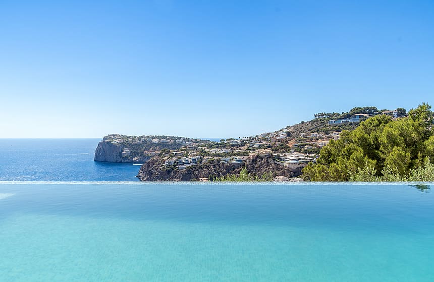 A clear blue infinity pool edge meets the horizon, overlooking a sunlit hillside with coastal villas and green trees.