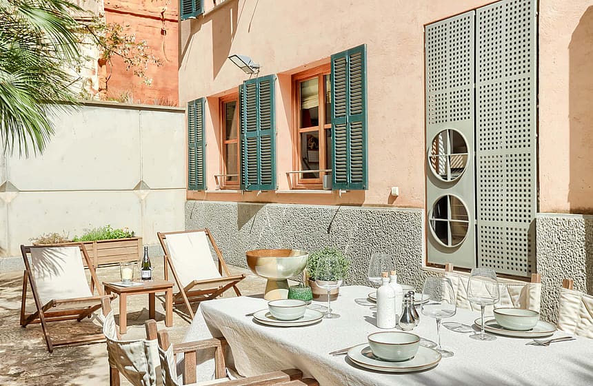 A private outdoor terrace in the Old Town with a dining table, folding wooden chairs, patterned stone wall, and green shutters.