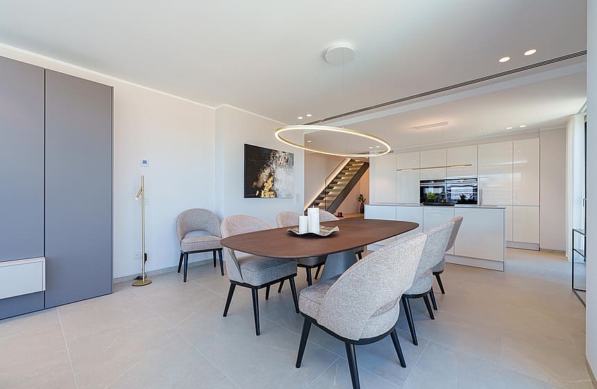 Comedor con mesa ovalada de madera y seis sillas, junto a una cocina blanca minimalista con iluminación empotrada.