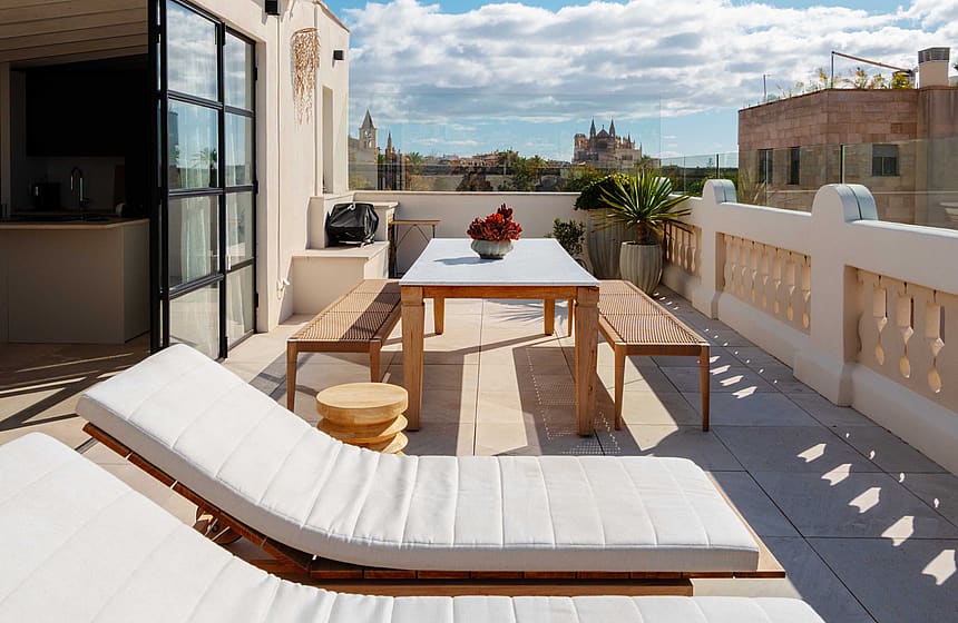 An expansive outdoor rooftop terrace featuring white sun loungers, a wooden dining table, and a view over the Santa Catalina skyline under a clear blue sky.