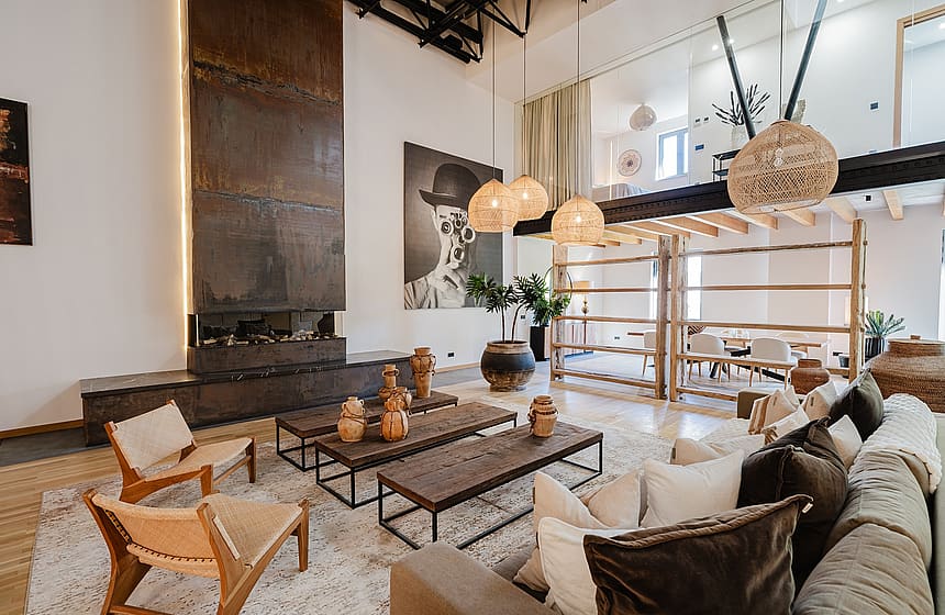 Modern open-plan living room with high ceilings, tall metal fireplace, wooden shelving, and warm pendant lighting.