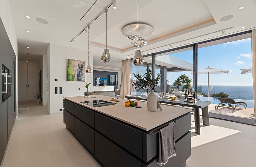 Modern open-plan kitchen with a large dark charcoal island, light stone countertop, and floor-to-ceiling glass doors opening to a sea-view terrace.