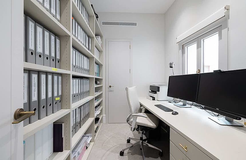 A compact home office featuring floor-to-ceiling built-in shelving packed with grey binders, a long desk with dual monitors, and a white task chair.