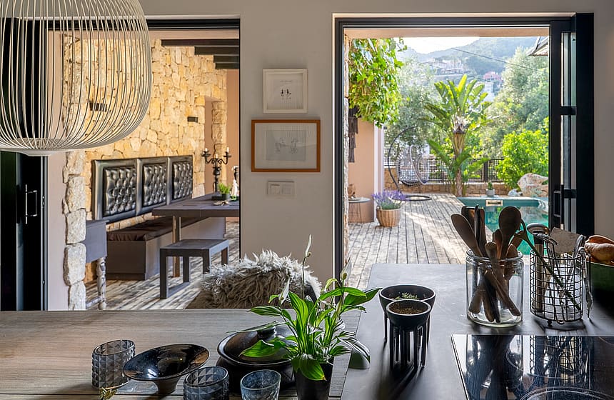 View from a kitchen counter looking out through open glass doors to a stone courtyard with green trees and mountain backdrop.